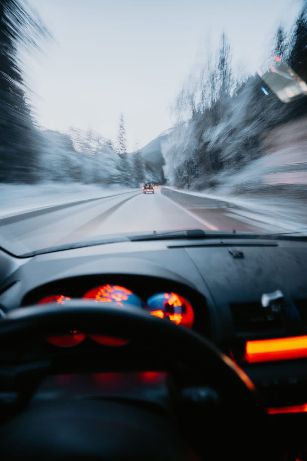 The view through a car windscreen on a snow-covered mountain road, another vehicle ahead and forest either side